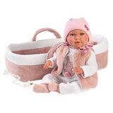 A realistic baby doll with a pink knit hat and bow, white outfit, and pink bunny vest, posed in front of a pink and white bassinet on a white background.