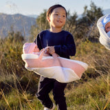 A young child stands outdoors in a grassy field carrying a baby in a soft pink and white bassinet or wrap. Another baby is partially visible on the right, and mountains and trees are in the background under a blue sky.