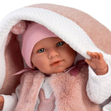 A close-up of a realistic baby doll with blue eyes, wearing a pink knit hat and a fluffy pink and white outfit, wrapped in a soft pink and white blanket.