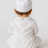 A baby wearing a white long-sleeve footed romper and matching white skullcap, photographed from behind against a light gray studio background.