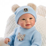 A baby doll or posed infant wearing a light blue knit beanie with two faux-fur pom-poms and a small square brand patch, plus a matching light blue knit top with a fox face applique, seated against a white textured backdrop.