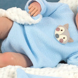 A close-up of a baby wearing a light blue knit romper with a gray and pink fox face patch on the chest, resting on a white textured blanket.