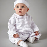 A baby wearing a white long-sleeve button-front romper, matching white bonnet, and white socks, seated against a plain gray studio background.