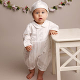 A baby wearing a white long-sleeve romper and matching bonnet stands barefoot next to a white table in a studio setting with a floral garland in the background.
