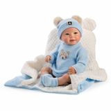 A baby doll wearing a light blue knit outfit with a matching beanie and booties, seated on a white textured blanket with blue trim against a plain white background.