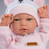 A close-up of a baby doll or toy wearing a pink knit outfit and a light pink fuzzy beanie with a white bear face patch. The beanie has pink pom-poms, and the outfit has a small tan brand patch reading 'Guca'.
