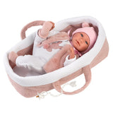 A realistic baby doll dressed in a white and pink outfit with a matching pink bonnet, lying inside a pink and white bassinet on a plain white background.