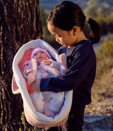 A baby in a pink bunny costume and hat is nestled inside a white and pink plush bunny-shaped bassinet, held by a young girl outdoors next to a tree.