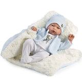 A baby doll or realistic baby toy wrapped in a white textured blanket with light blue trim, wearing a white outfit, matching hat, and a knitted blue and white cardigan, photographed on a white background.