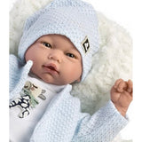 A realistic reborn baby doll wearing a light blue textured knit bonnet and matching waffle-knit cardigan over a white top, posed on a white faux-fur surface against a white background.