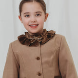 A young girl with her hair pulled back, smiling at the camera, wearing a brown coat with a large ruffled velvet collar and bow.